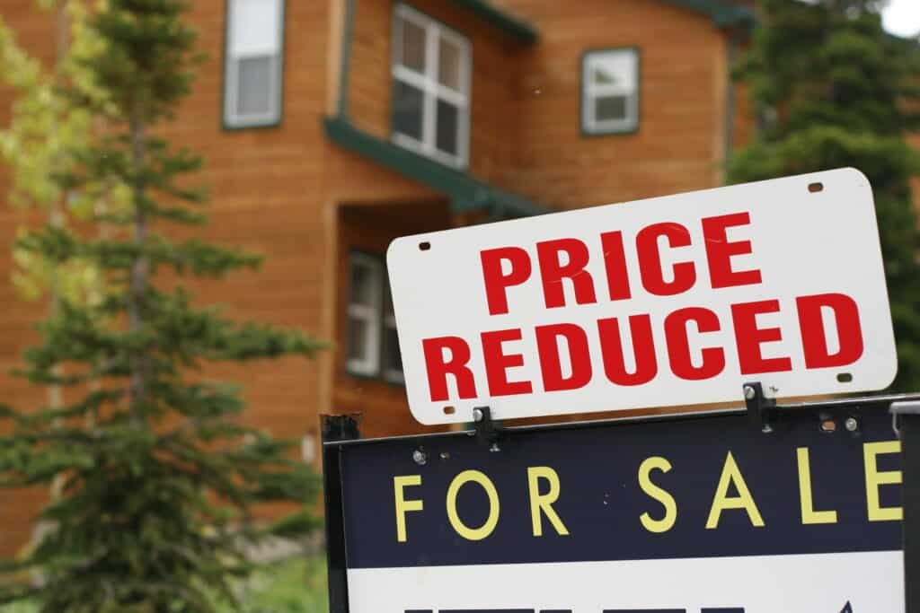 A for sale sign outside of a home with a price reduction sign rider on top.