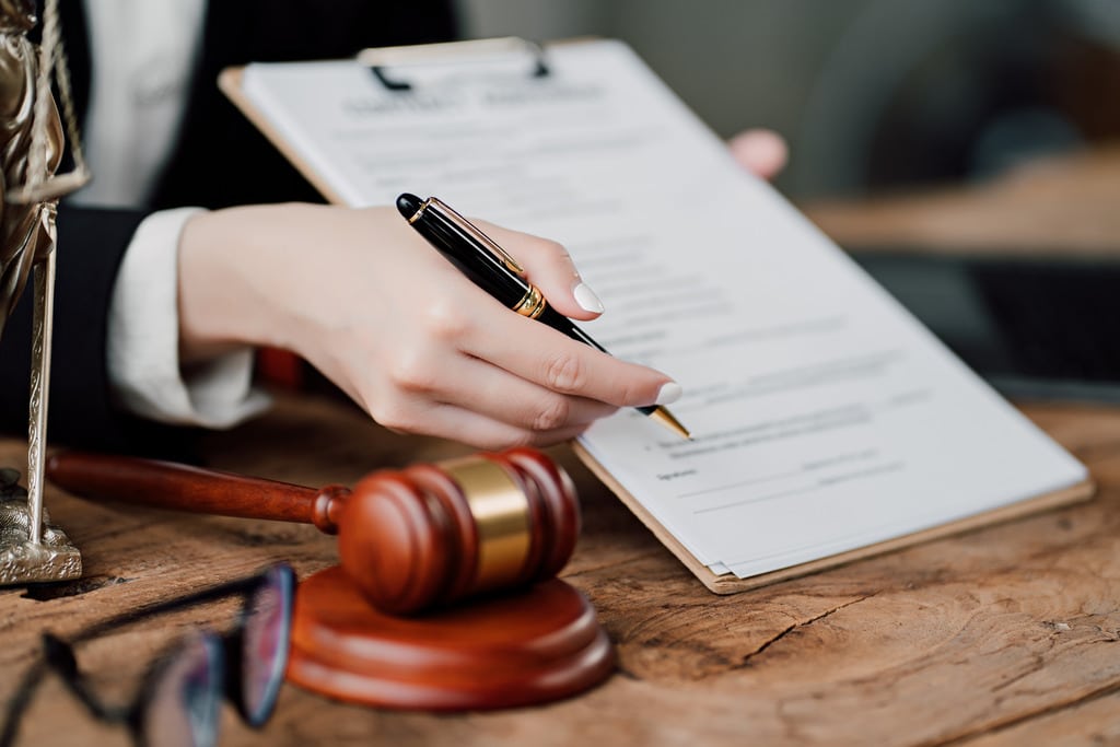 A hand holding a pen pointing out where to sign a legal document. There is also a gavel on the left.