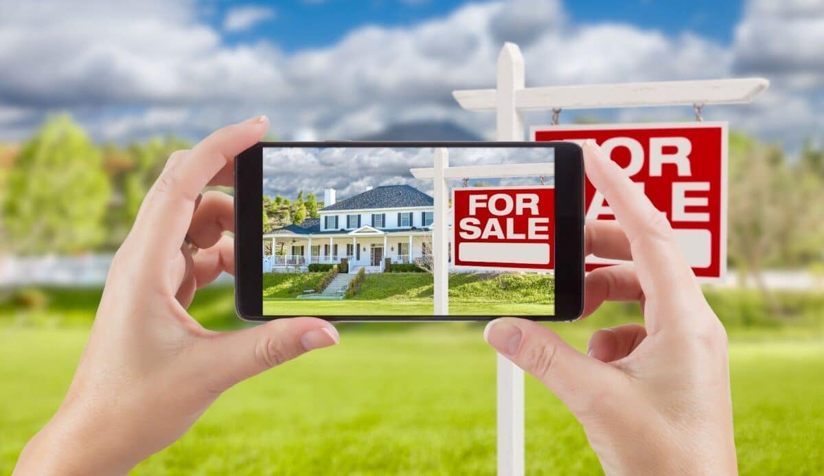 A person taking a photo of a house with a For Sale sign in front.