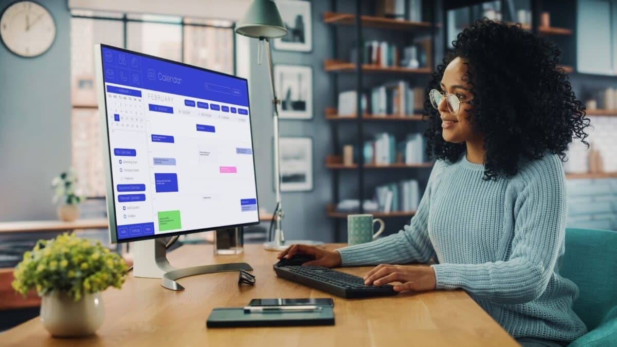  A woman sitting at a desk looking at the computer with a calendar showing scheduled events on it.