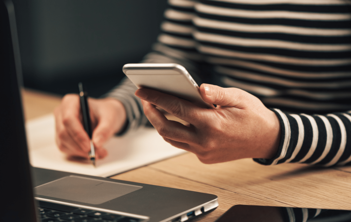 Person sitting at a laptop looking at a mobile phone and taking notes.