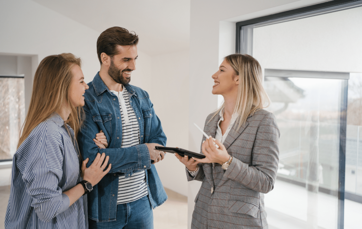 A young female real estate agent is holding a tablet and talking with a young couple inside a house.