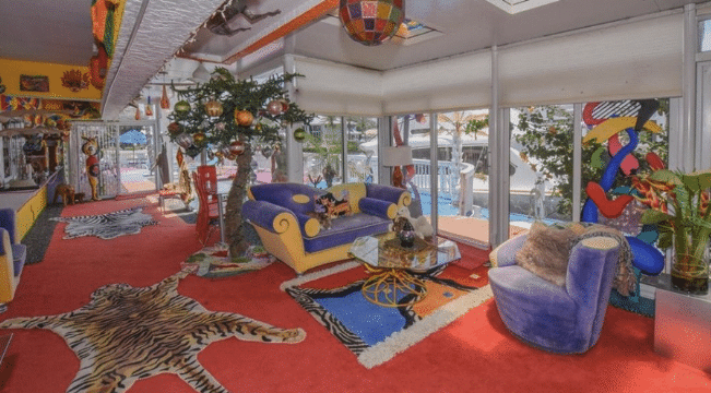 A large sitting room with all glass exterior floor-to-ceiling windows, red carpeting, a tiger skin rug, a zebra skin rug, and a purple and yellow sofa across from a purple velvet chair.