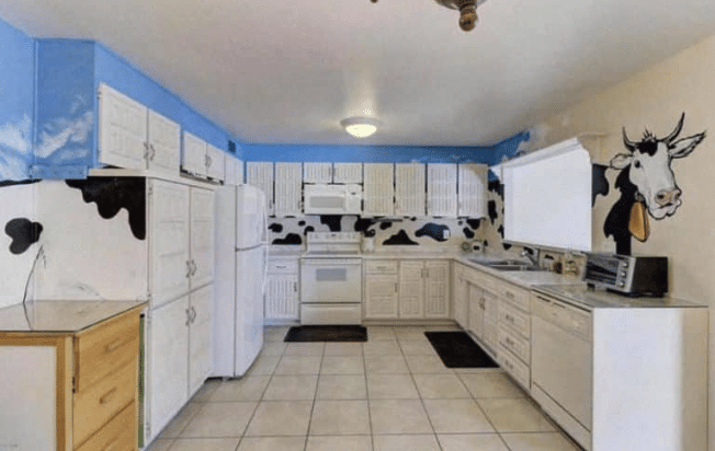 U-shaped kitchen with white appliances and counters and a black and white cow painted from one end to the opposite end with blue sky.