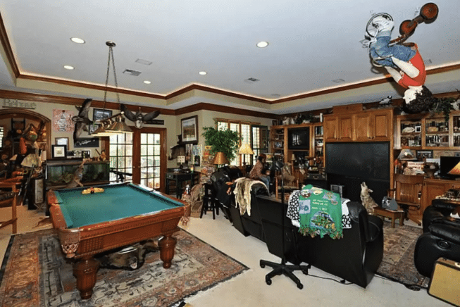 Entertainment room with a large pool table, a sofa, a large TV, lots of knick-knacks, more shelving, and a mannequin of a young boy riding a tricycle hanging upside down from the ceiling.