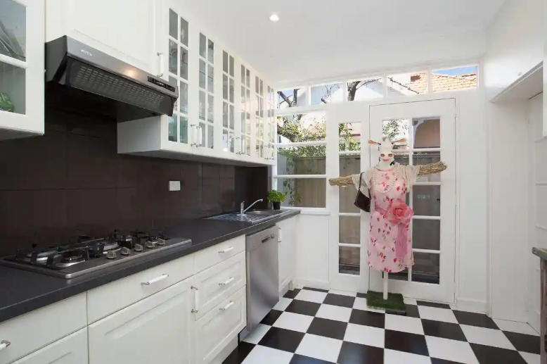 Black and white kitchen with black and white alternating tiles on the floor and a well-dressed cow scarecrow standing at the opposite end of the kitchen, arms outstretched.