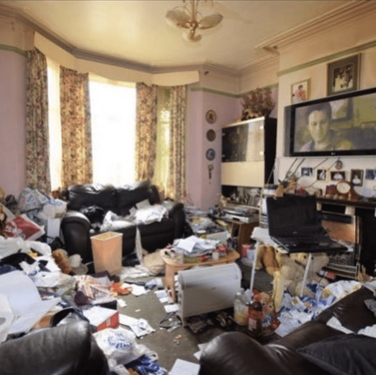 Living room with light pink and avocado walls and floral print drapes. There are a couple of chairs and a sofa situated in front of a large TV on the wall. The whole room is covered in trash to the point it's difficult to see the floor in many places.