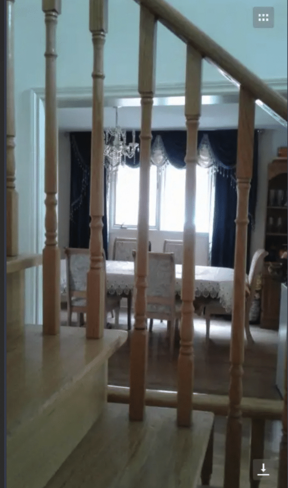 Dining room with six chairs, a large window with scalloped drapes, from the point of view of the staircase. The stair railing is prominent in front of the dining room.