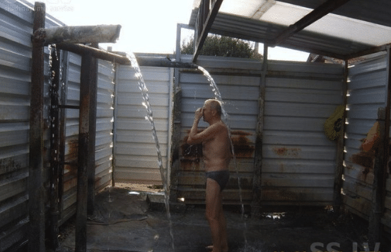 Elderly man in his underpants standing in a metal enclosure where water falls from two tubes. He's standing under the water.