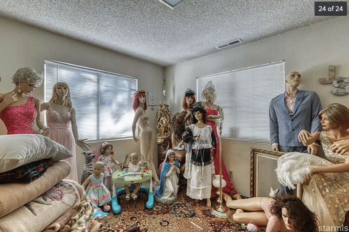 Room with two large windows and eight mannequins standing around the room in various stages of dress. One mannequin is lying on the floor, almost crawling. There are also several small child-size mannequins in various poses and a Virgin Mary kneeling in front of a cluster of mannequins.