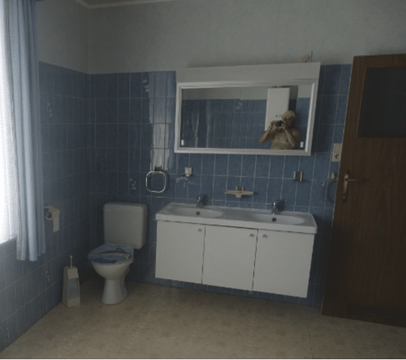 Blue-tiled bathroom with a mirrored medicine cabinet above a double vanity sink and a toilet to the side. In the mirror is a shirtless man's reflection as he's taking this photo.