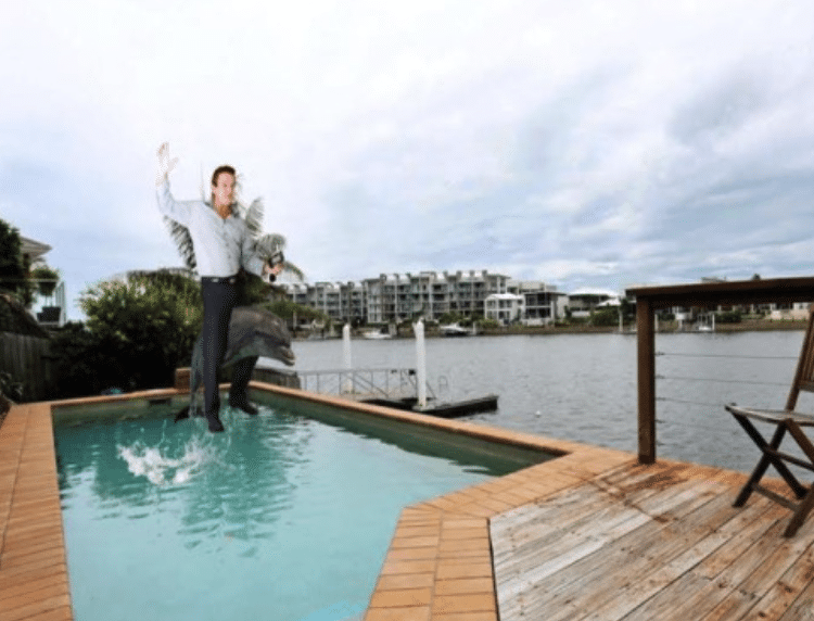 A small pool outside a waterfront property with a man wearing slacks and a button shirt superimposed on the back of a dolphin jumping out of the pool.